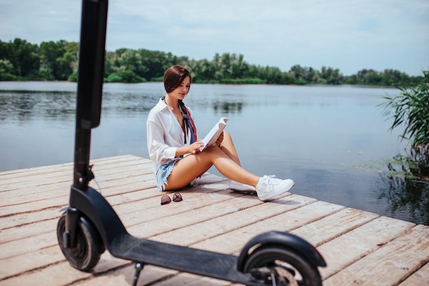 A beautiful girl with an electric scooter reads a book on a wooden bridge by the river. eco transport and health care concept.