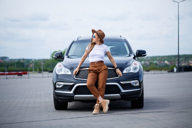 A beautiful girl with glasses and a brown hat is standing near a black car. Young woman with car in the parking lot