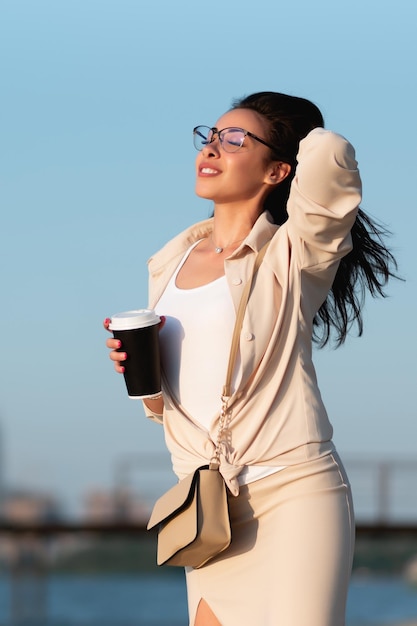 Foto bella ragazza con gli occhiali si trova sulla spiaggia vicino al fiume gode di una passeggiata tiene un bicchiere di carta con una bevanda calda in mano mockup di caffè