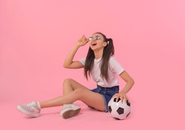 Красивая девушка со счастливой улыбкой сидит на полу с мячом на розовом фоне