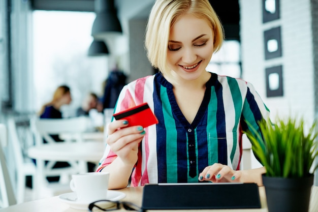Beautiful girl with light hair wearing colorful shirt sitting in cafe with tablet, credit card and cup of coffee, freelance concept, online shopping.