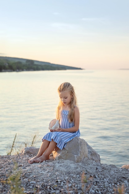 A beautiful girl with long blond hair sits on the shore and admires a large sea shell