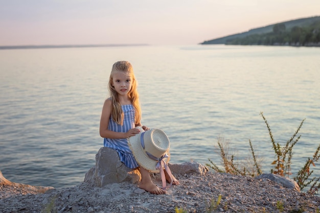 Beautiful girl with long blond hair sits on a stone on the banks of a river lake sea