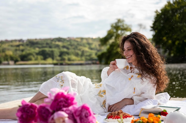 A beautiful girl with long curly hair dressed in a white dress serving cup of a tea have picnic in a sunny time