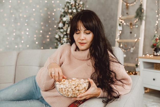 長い黒髪の美しい少女がソファに横になり、クリスマスツリーの背景にポップコーンを食べます。