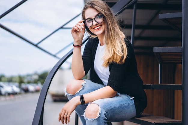 Beautiful girl with long hair and glasses sitting on metal stairs on the wooden background of house with vertical boards