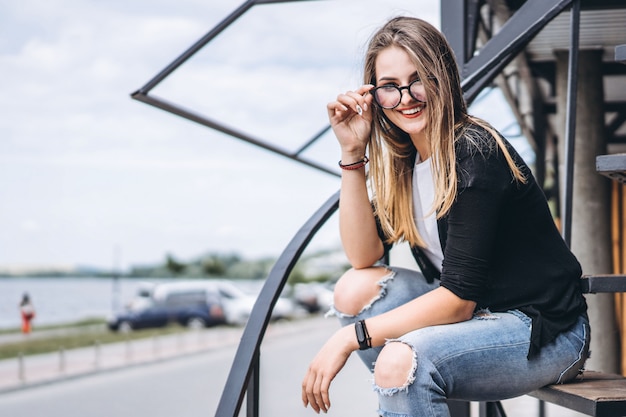 Beautiful girl with long hair and glasses sitting on metal stairs on the wooden background of house with vertical boards