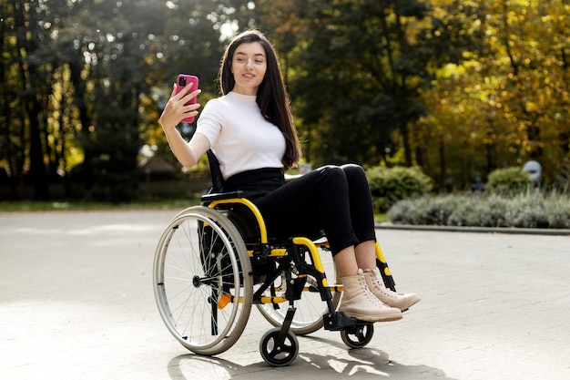 Beautiful girl with long hair sits in a wheelchair takes a selfie on a smartphone Beautiful young woman on the street