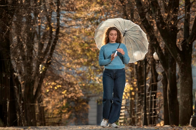Beautiful girl with long hair walks through autumn park Young woman in blue sweater with transparent umbrella in hands
