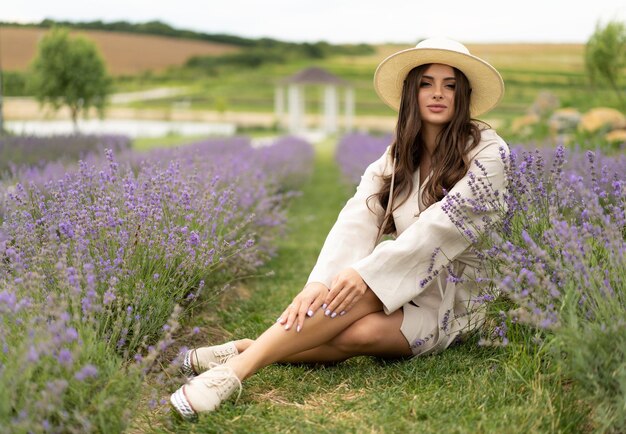 A beautiful girl with long hair and wearing a hat poses outdoors in a field of lavender