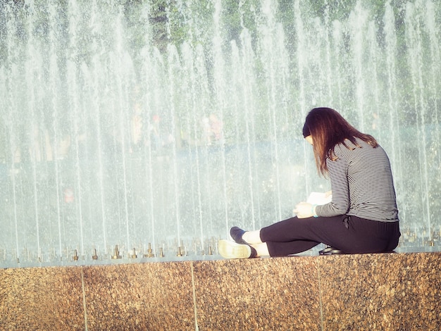 A beautiful girl with long natural black hair sits on the bokeh background of fountain. Autumn euphoria in faded tones. Back view.