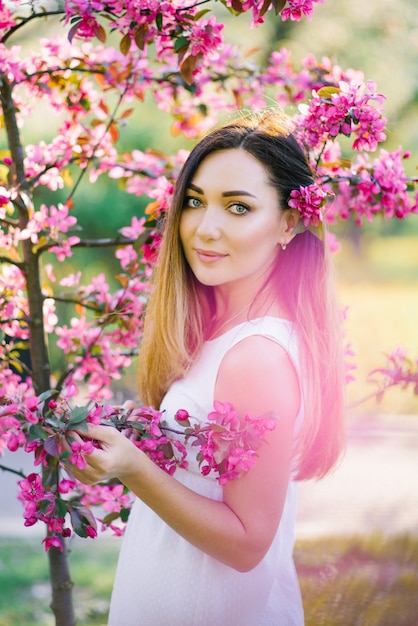 A beautiful girl with long straight hair and big eyes stands in the flowers of a pink Apple tree. Professional makeup