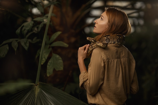 Красивая девушка с естественным макияжем и рыжими волосами стоит в джунглях среди экзотических растений со змеей.