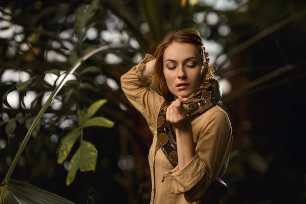 A beautiful girl with natural make-up and red hair stands in the jungle among exotic plants with snake.