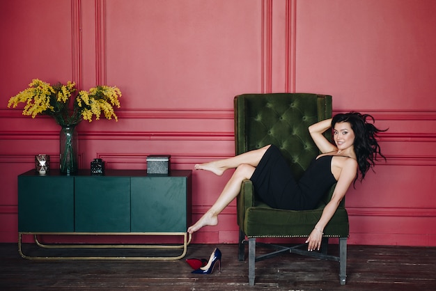 Beautiful girl with oriental appearance. Black fitting dress on the straps. Red wall. Green chair.