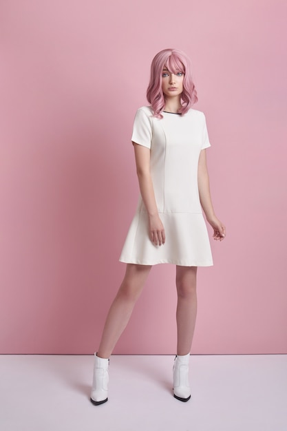 Beautiful girl with pink hair, hair coloring. Cute anime woman stands on a pink background in a short white dress