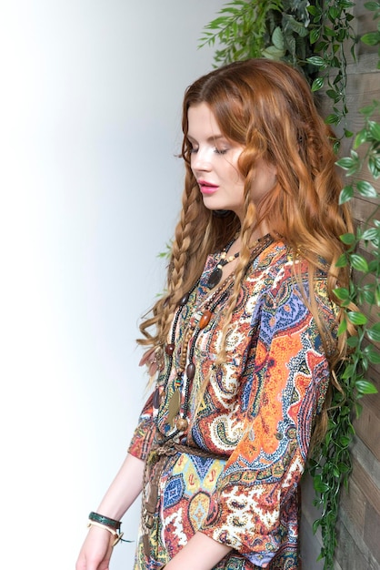 A beautiful girl with red curly hair stands on the corner of a white and wooden wall