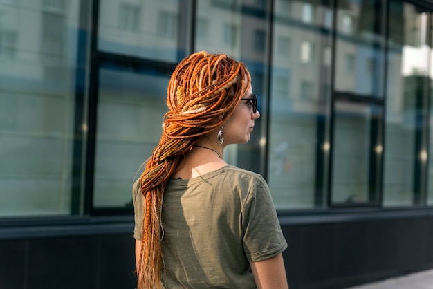 Beautiful girl with red dreadlocks poses against the background of an office building. Street photo