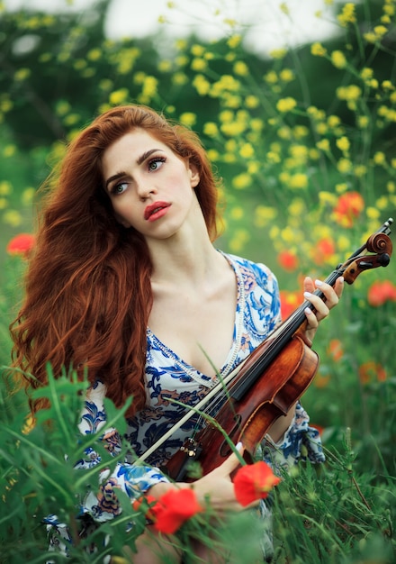 Beautiful girl with red hair and blue dress holding violin on nature field of flowers.