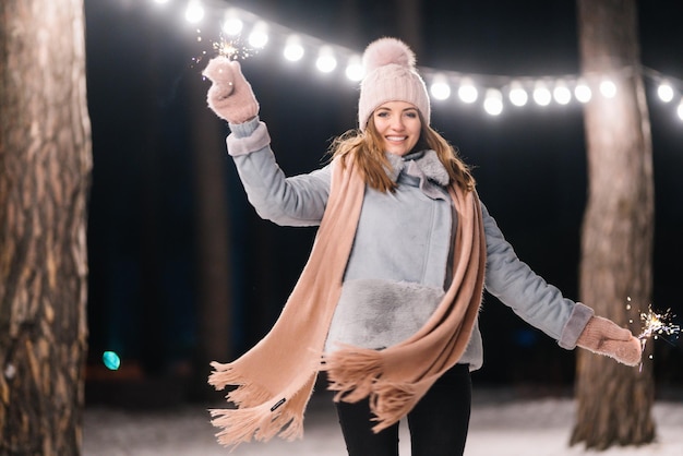 Beautiful girl with sparklers in hands Happy winter time in forest Festive garland lights Christmas