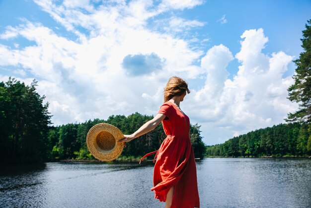 湖の近くの麦わら帽子を持つ美しい少女。素晴らしい夏の自然。旅行のコンセプト。ワンダーラストのインスピレーション。リネンの赤いドレスを着た女性。衣類の環境にやさしい素材。