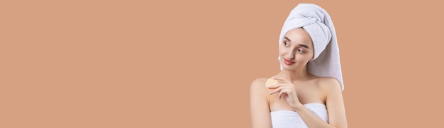 Beautiful girl with thick eyebrows and perfect skin at calming\
coral background towel on head beauty photo holds a cosmetic\
sponge