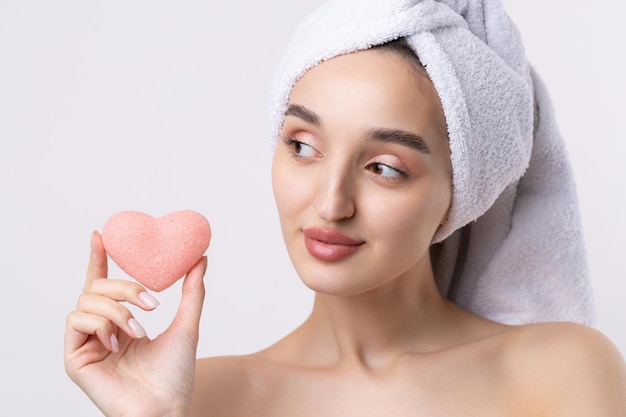 Beautiful girl with thick eyebrows and perfect skin at white background towel on head beauty photo Holding a cosmetic pink heart sponge