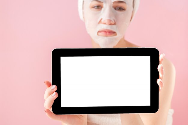 Beautiful girl with a tissue mask and a tablet in their hands with copy space on a pink background.