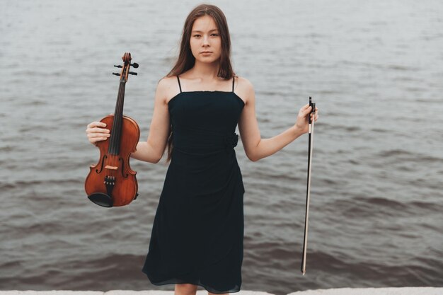 A beautiful girl with a violin stands against the background of water. Asian appearance. musical concept. High quality photo