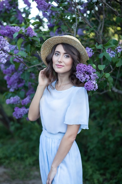 beautiful girl with wavy brown hair in a straw hat in a lilac garden in bloom summer vacation
