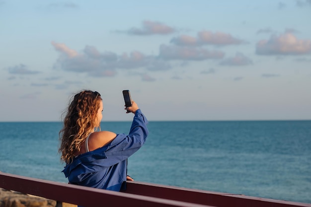 A beautiful girl with wavy hair stands on the bridge and takes a sea sunset on the phone