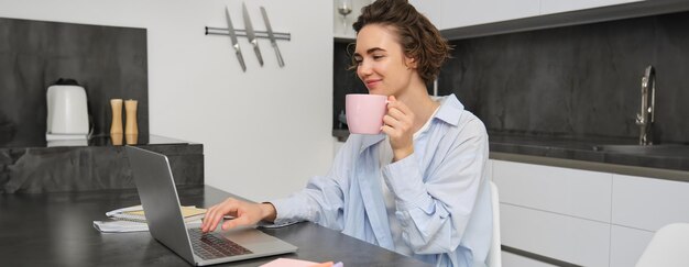 Photo beautiful girl working from home drinking coffee and looking at laptop young businesswoman sits in