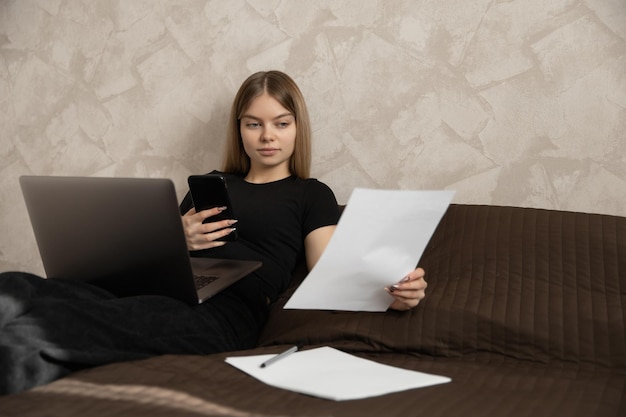 Beautiful girl works at home at the computer lying on the bed\
and checks the papers