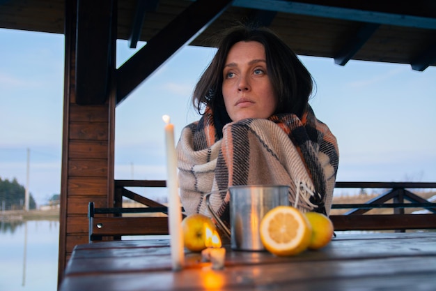 A beautiful girl wrapped in a plaid sits at a table in a street gazebo with a metal mug of tea