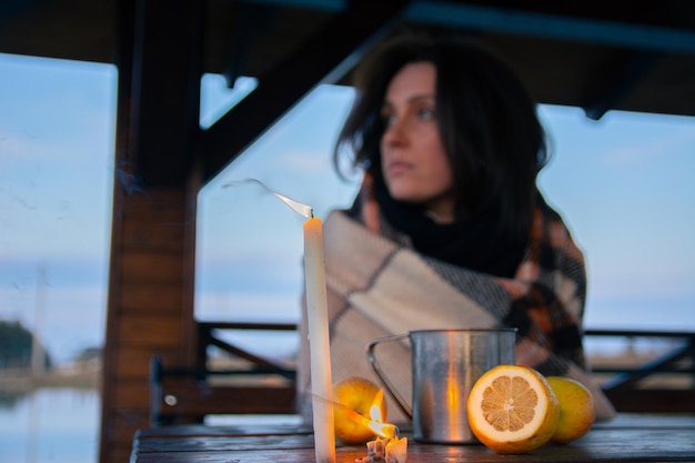 Una bella ragazza avvolta in un plaid siede a un tavolo in un gazebo di strada con una tazza di tè di metallo