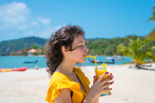 Beautiful girl in a yellow dress drinks fresh mango on the beach of a paradise island. Perfect vacation.