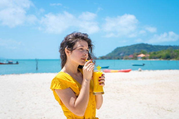 Beautiful girl in a yellow dress drinks fresh mango on the beach of a paradise island. Perfect vacation.