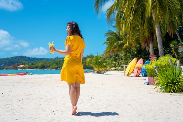 La bella ragazza in un vestito giallo beve il mango fresco sulla spiaggia di un'isola di paradiso. vacanza perfetta.