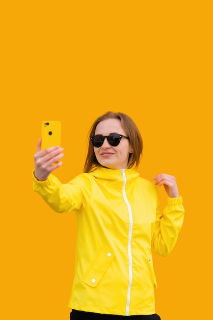 A beautiful girl in a yellow jacket takes a selfie on orange background