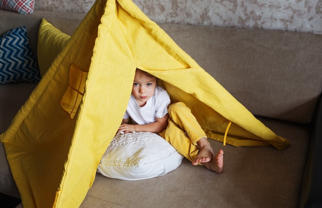 A beautiful girl in yellow pants and a white T-shirt plays with the inside of a teepee at home on the couch. Home games for children