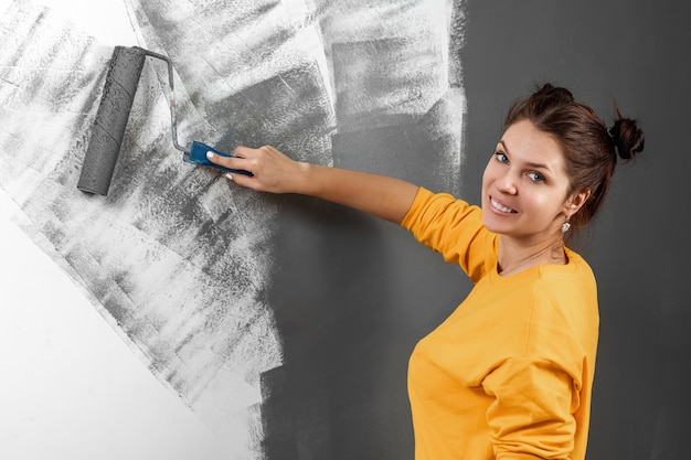 Beautiful girl in a yellow sweater paints a wall in gray paint. Room painting, repair, design. Copy space.