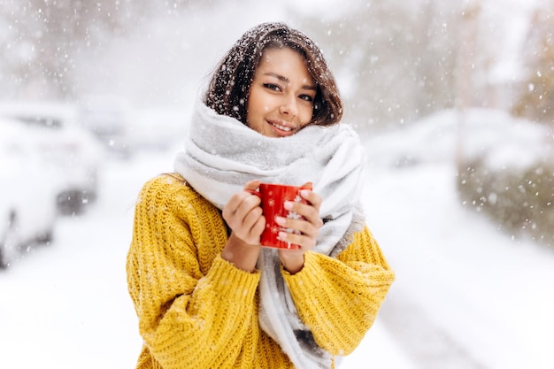 Beautiful girl in a yellow sweater and a white scarf standing with a red mug on a snowy street