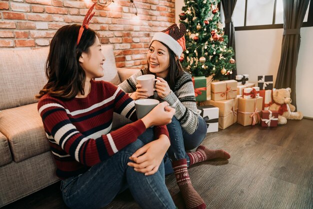 beautiful girlfriends christmas celebration enjoy mulled wine at home. saying merry christmas and happy new year to each other. young girls sitting on the wooden floor indoor in winter.