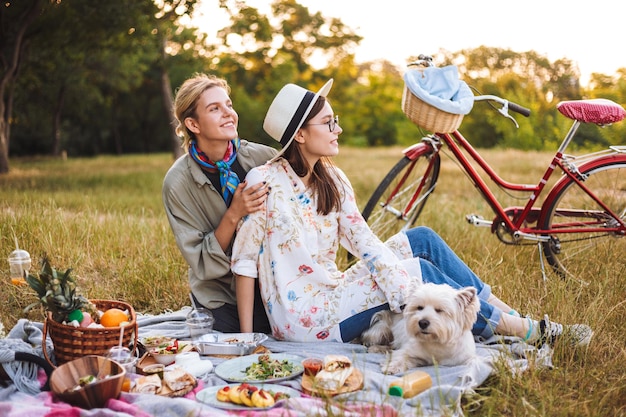 Beautiful girls dreamily looking aside spending time on picnic with cute little dog in park with red bicycle on background