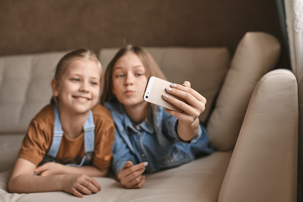 Beautiful girls take selfies on their phone while lying on sofa. Sisters take a break from homework and take photos during quarantine