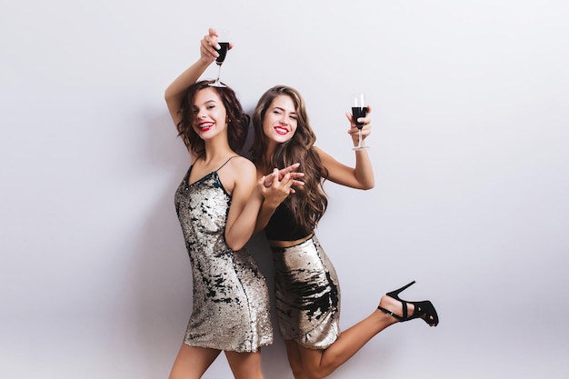 Beautiful girls, two friends smiling and raising glass of red wine, having fun. Wearing dresses with sequins, girl has leg up with high heels sandal. Sexy, stylish look. Isolated