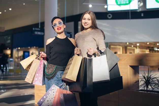 Beautiful girls with shopping bags are looking at camera and smiling while doing shopping in mall