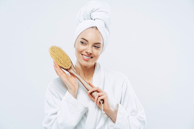 Beautiful glad woman with healthy skin and toothy smile holds wooden massage brush