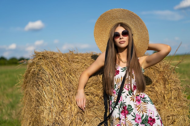 Beautiful glamorous girl in a dress and a straw hat on a field near a haystack