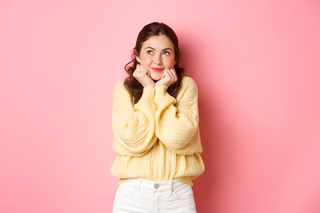 Beautiful glamour girl thinking about love looking up and daydreaming imaging something cute and romantic standing against pink wall